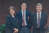 A young man stands besides his older parents for a photo, all three dressed formally.