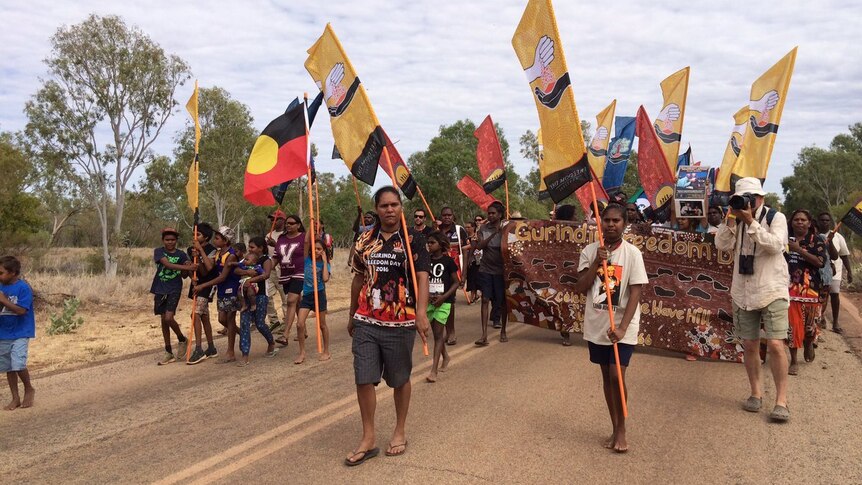 A group of Aboriginal people march with flags along a road in Kalkarindji