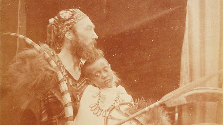 Old black and white portrait of a Victorian man draped in lion fur and holding a young African boy on his lap who looks sad