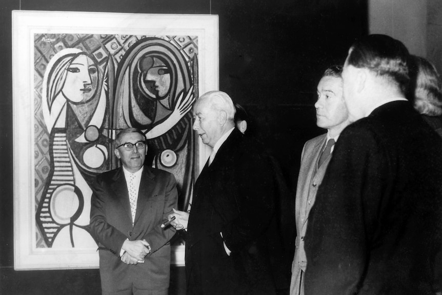 Black and white archival image showing group of older men standing in front of a modernist painting by Picasso.