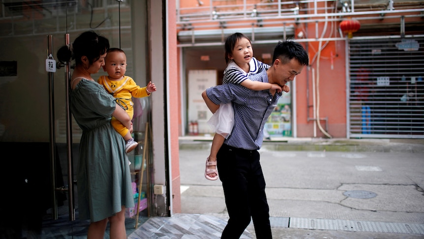 A man and a woman each carry a child as they leave a building. 