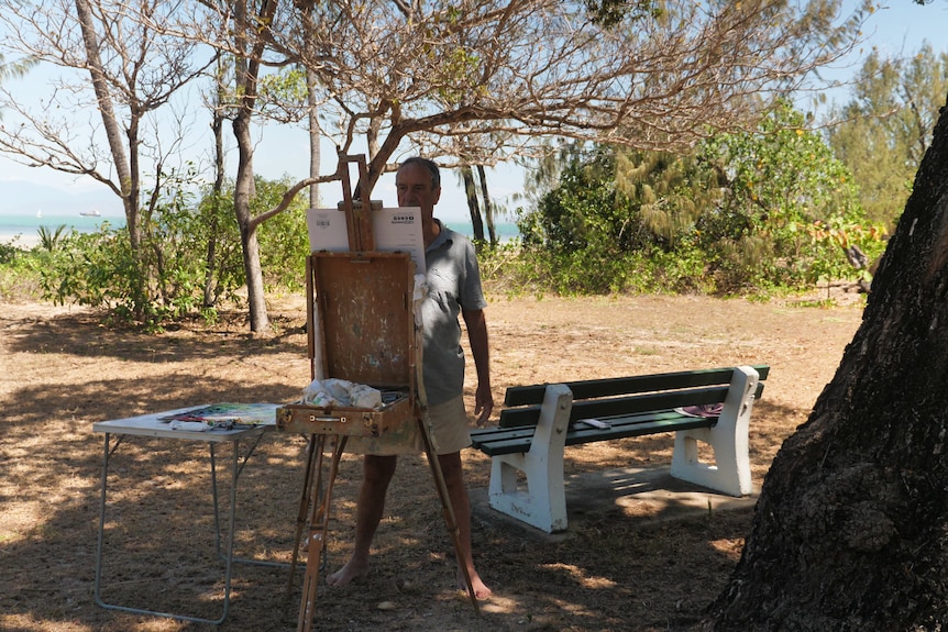 a man paints on an easel in a bush park next to a beach