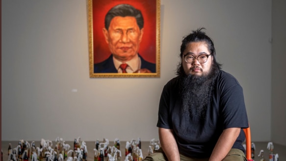 Artist Badiucao sits on a chair in front of a painted picture of Xi Jinping. There are recycled glass bottles on the floor 