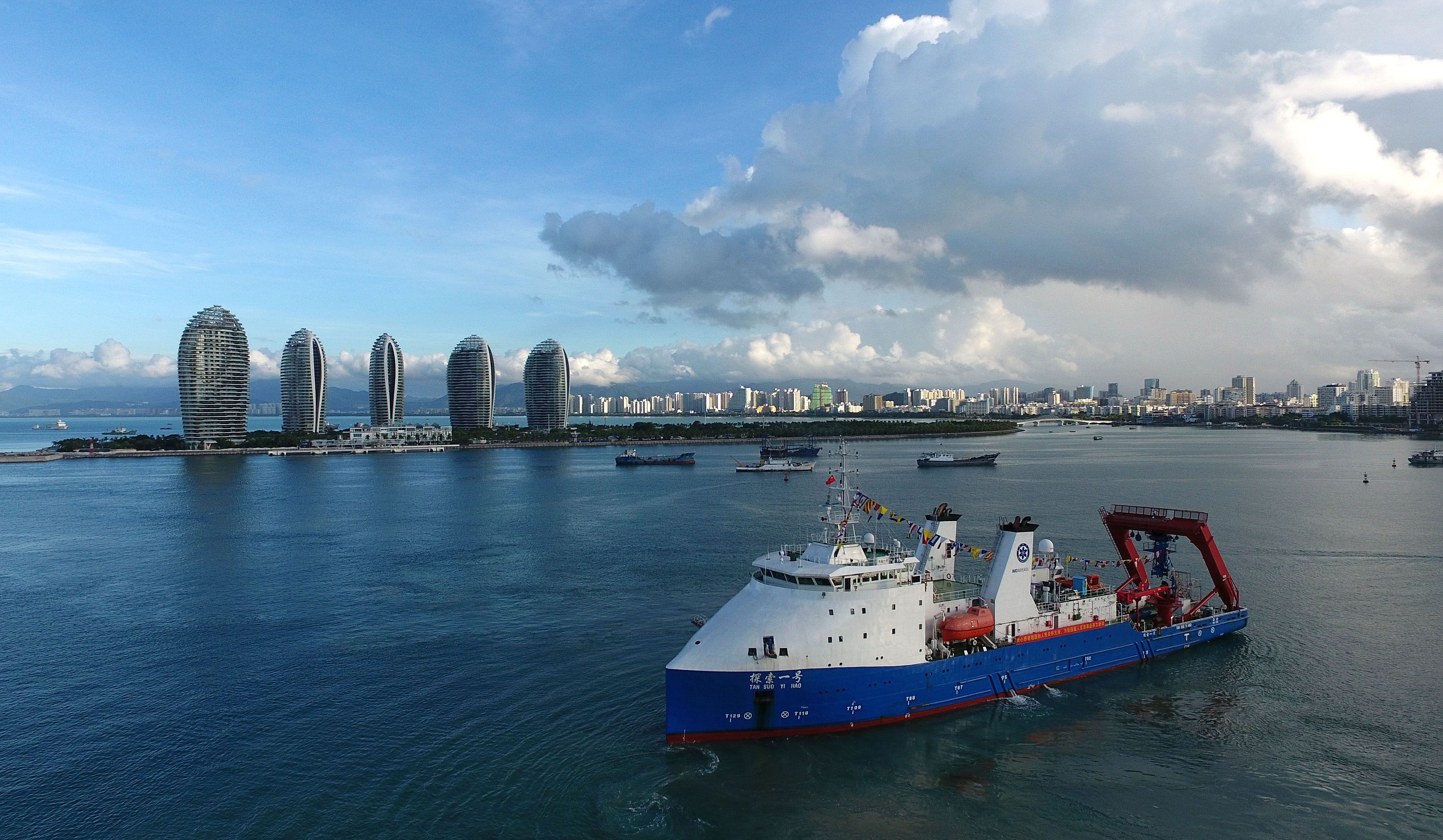 A large ship with white cabin at front, blue bottom and large flat back deck in port in front of large city.