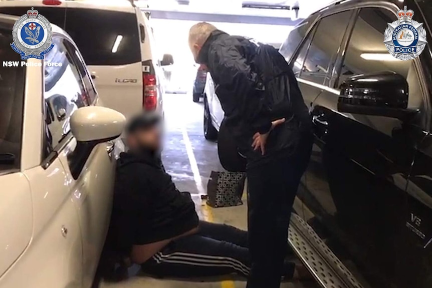 A man sits on the floor leaning against a car as a police officer stands over him.