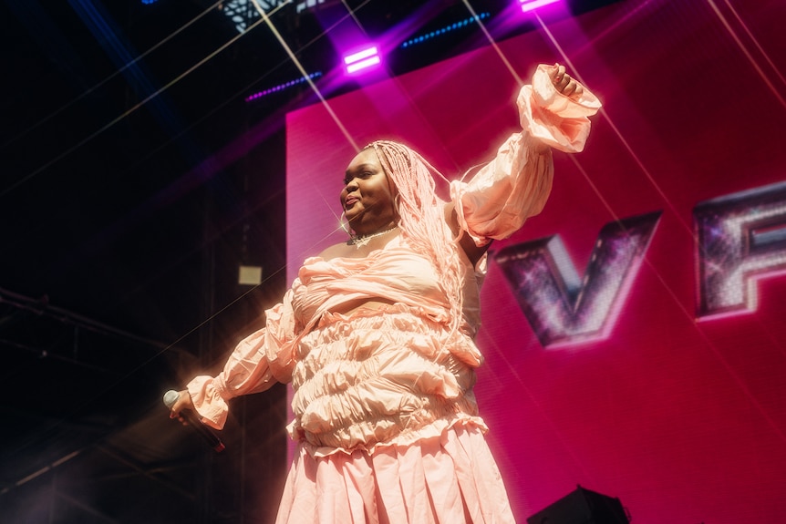 Vv Pete wearing pink, with pink braids holding her hands out on stage