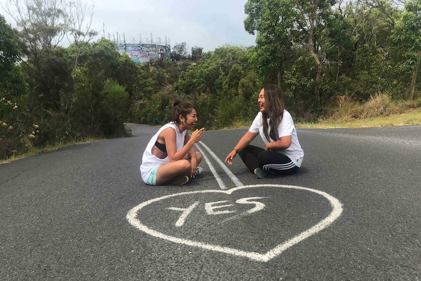 Two young women sit in the middle of a road laughing. In front of them the word 'yes' is spray painted inside a white love heart