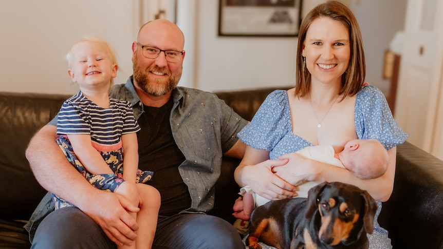 a husband and wife with their toddler and young baby, plus a sausage dog, smile at the camera in a family photo