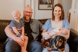 a husband and wife with their toddler and young baby, plus a sausage dog, smile at the camera in a family photo