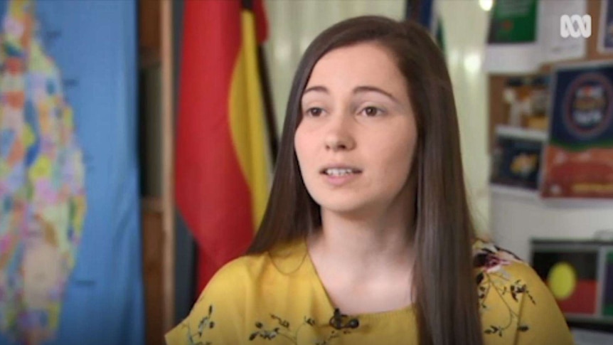 Head and shoulders image of a young woman speaking in a classroom environment with Indigenous flags hanging in the background.