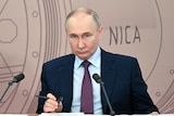 Vladimir Putin sits before microphones with the words NICA on the background 