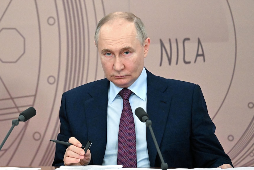 Vladimir Putin sits before microphones with the words NICA on the background 
