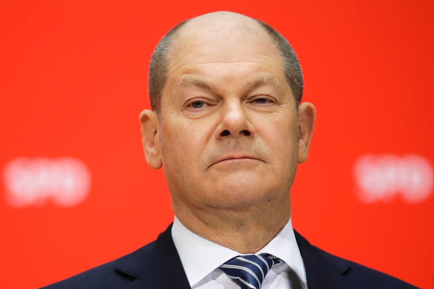 German Chancellor Olaf Scholz stands in front of red backdrop