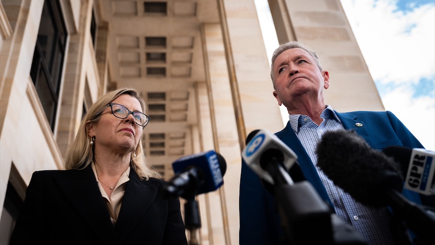 A smartly dressed man and woman stand in front of microphones, shot from low down.