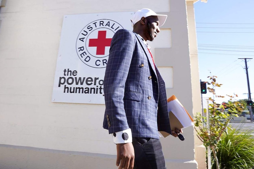A tall man in a suit jacket carrying some paperwork walks past a Red Cross sign in Mount Gambier.
