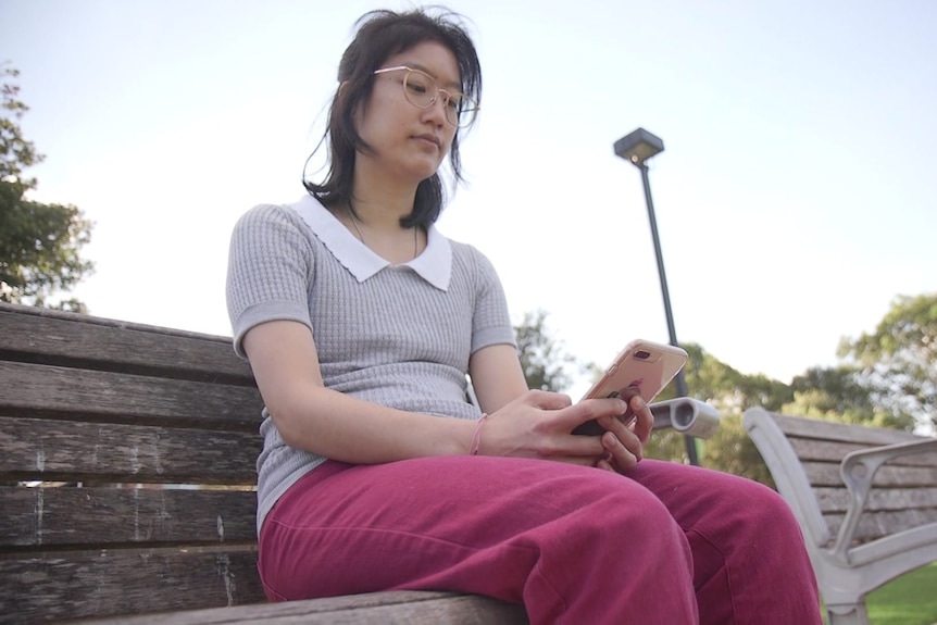 An Asian woman wearing a grey shirt and purple pants sits on a park bench.