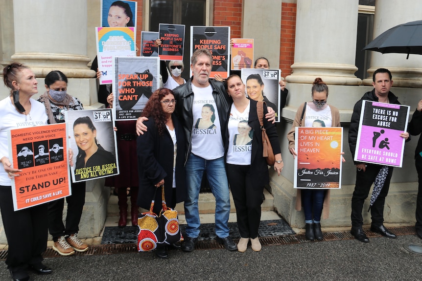 A group of people carrying posters and placards stands outside court posing for a photo with some with arms around each other.