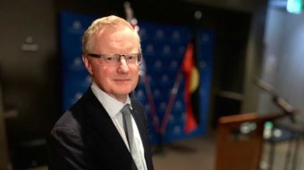 RBA Governor Philip Lowe stands in front of Australian and Aboriginal flags