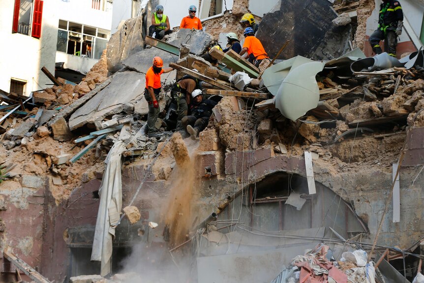 You view the rubble of a multi-storey masonry building with rescue workers in orange nestled between fragments as dust falls.