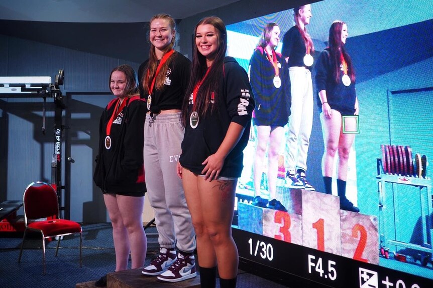 Three young women stand on a podium in front of a big screen with medals around their necks