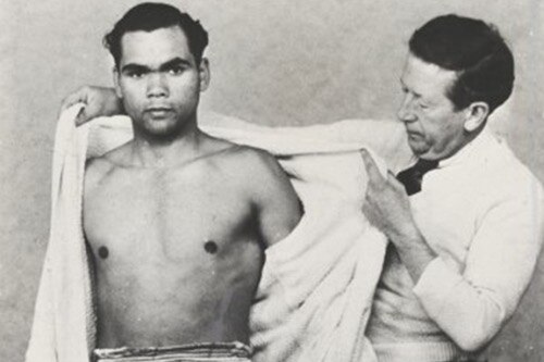 A black and white photo of an Indigenous boxer stands alongside his trainer who is taking the boxer's robe off.