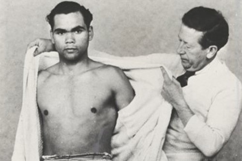 A black and white photo of an Indigenous boxer stands alongside his trainer who is taking the boxer's robe off.