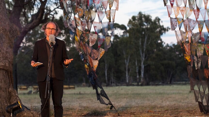 A man in a black suit talking into a microphone in front of coloured flags hung from a tree.