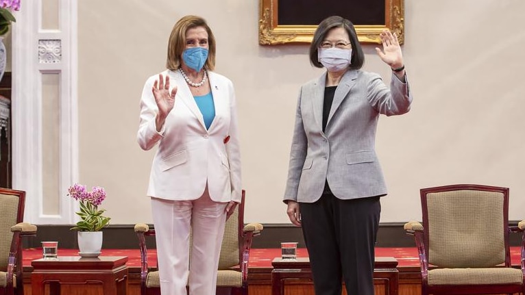 Two women, one in a white pant suit and the other in a grey and black pant suit, wave