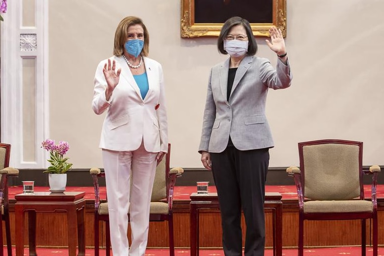Two women, one in a white pant suit and the other in a grey and black pant suit, wave
