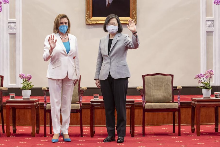 Two women, one in a white pant suit and the other in a grey and black pant suit, wave