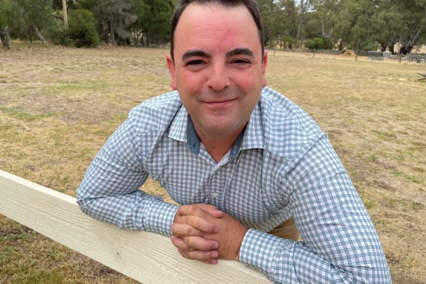 A smiling young man in a blue-check shirt leans on a fence in a paddock.