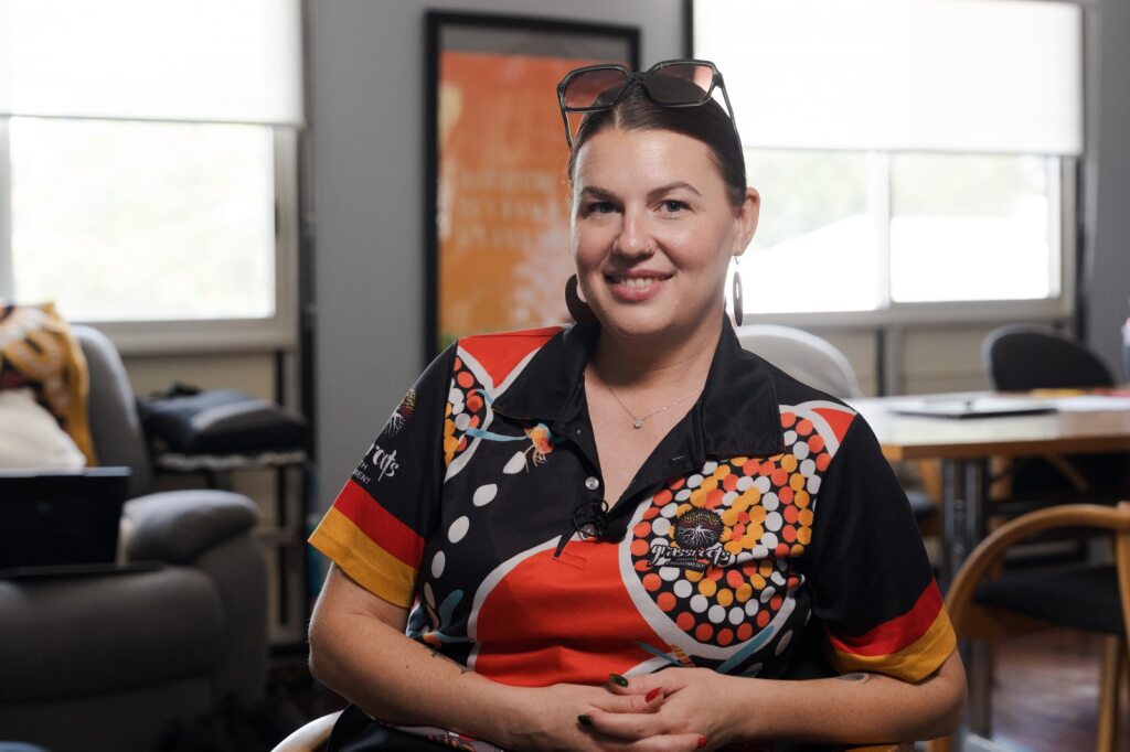 Uma mulher vestindo uma camisa pólo com estampas indígenas e óculos na cabeça enquanto sorri. 