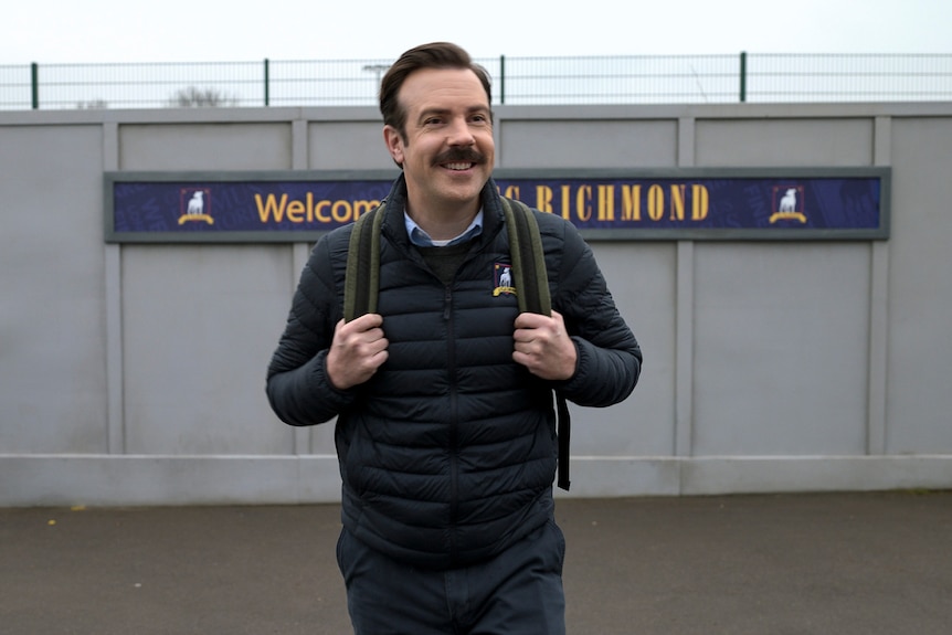 A 40-something man with a moustache and a backpack grins. Behind him is the wall of a soccer stadium. 