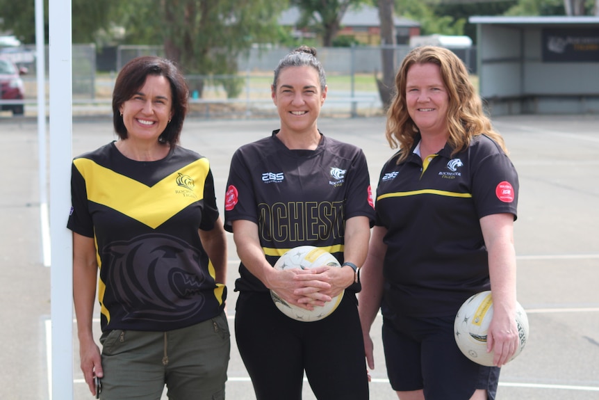Three women stand on a bitumen netball court and smile.