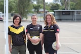 Three women stand on a bitumen netball court and smile.
