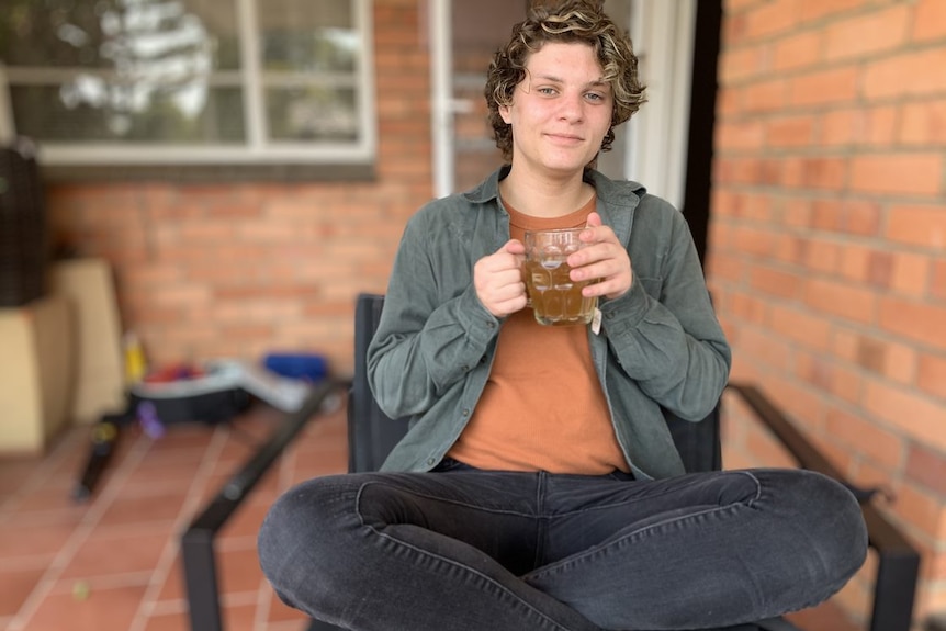 A young man holding a cup of tea smiles at the camera.