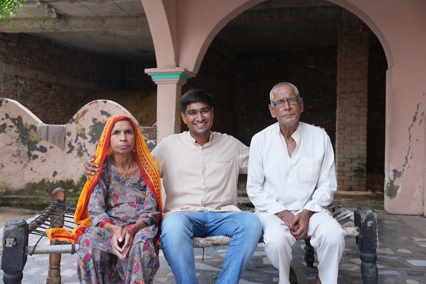 Ramveer Tanwar sits between two older people, smiling, with his arms around their shoulders.