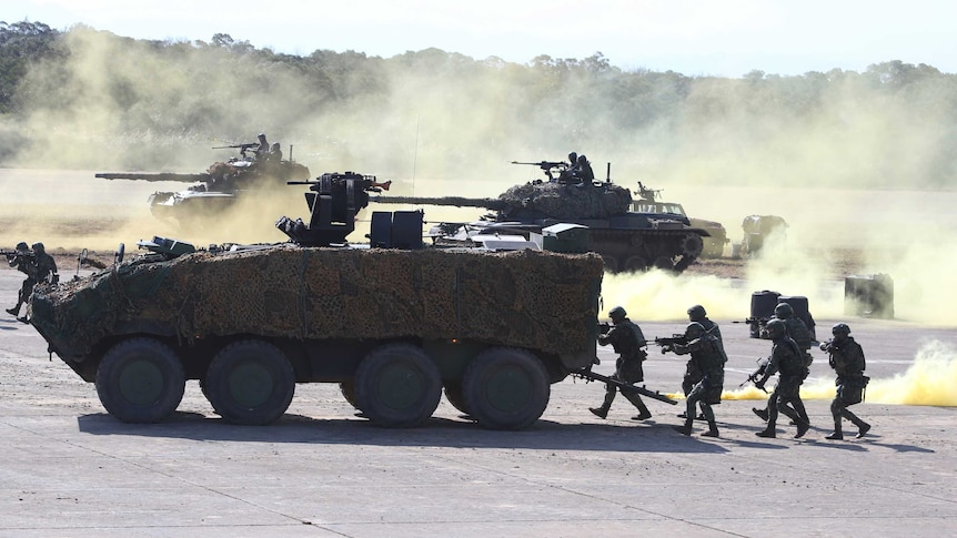 Taiwanese soldiers take part in a military drill. Troops move behind a camouflaged vehicle, there are tanks in the background.