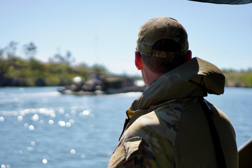 A back of male soldier's head as he looks out over the water. He is wearing a cap and a khaki jacket.