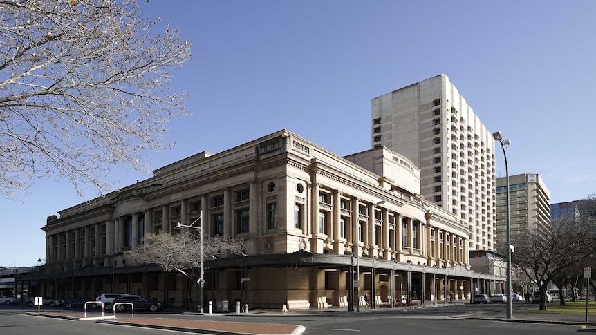 A grand building with columns with a tall building behind and a tree with blossoms in the foreground