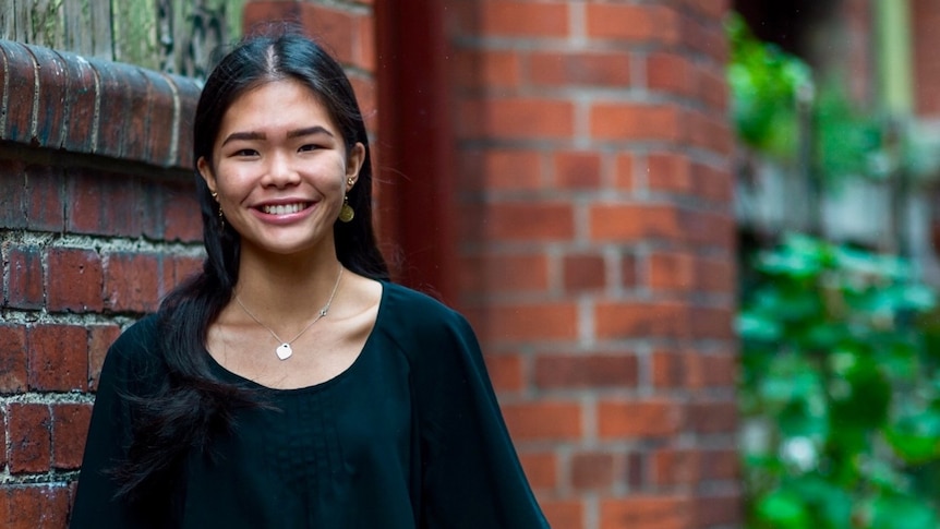 A young woman with dark hair and a black tshirt stands in front of brick house, smiling