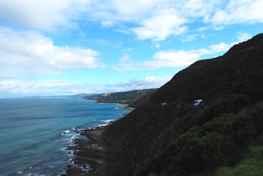 A wide shot of a bus rounding a bend on a windy coastal road. The ocean and green pastures in the distance.