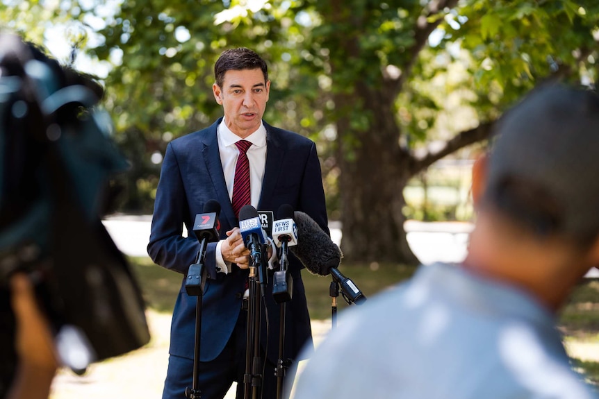 A man in a blue suit and a red tie speaks to the media while standing in a park.