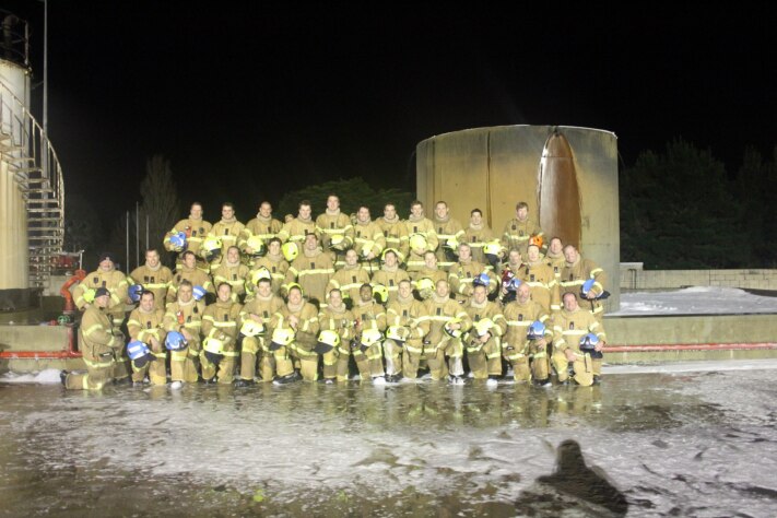 A group photo of 38 firefighters in yellow uniforms and holding helmets, stand for a group photo at training centre outdoors