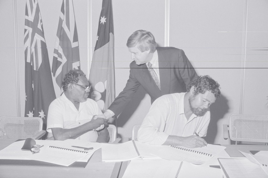 A black and white photo of an Indigenous man and a white man shaking hands, while another man signs a document.