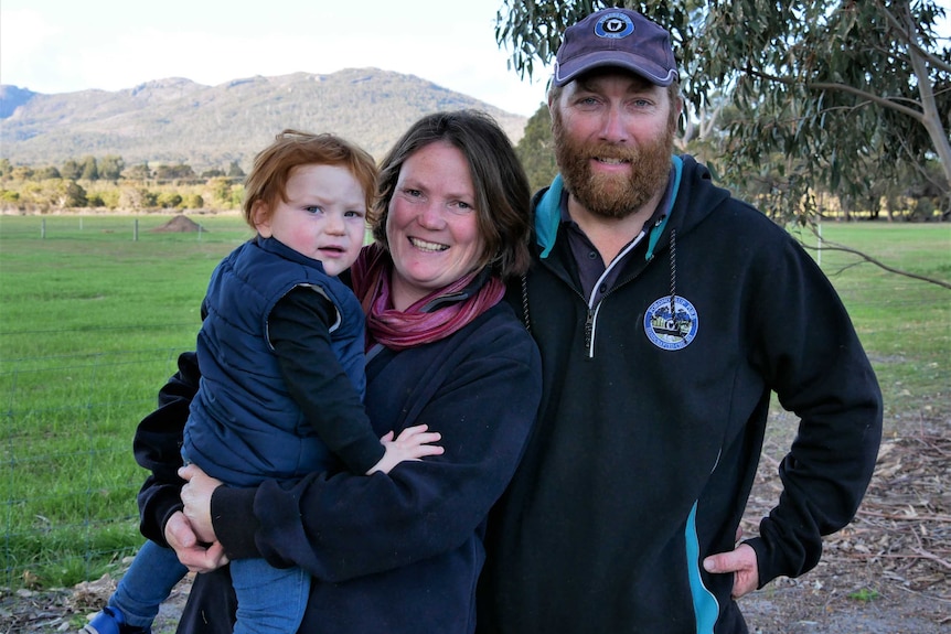 A man, woman and young child stand in a paddock in front of a large hill.