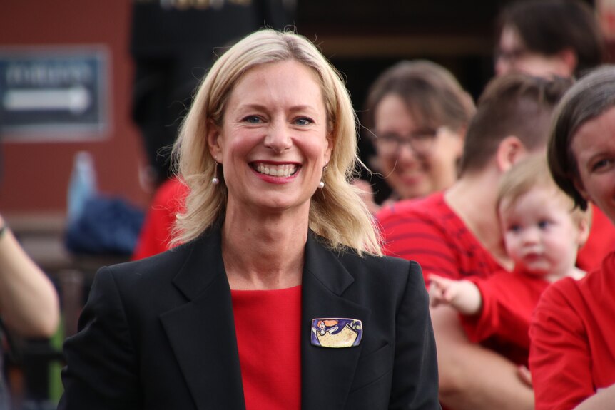 Rebecca White, wearing a red top and black jacket, smiles at the camera