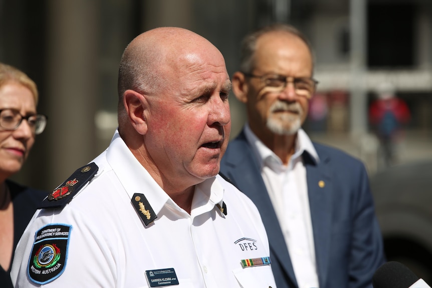 A middle-aged man in a DFES uniform speaks to reporters off-frame at a press conference. Man and woman blurry in background.