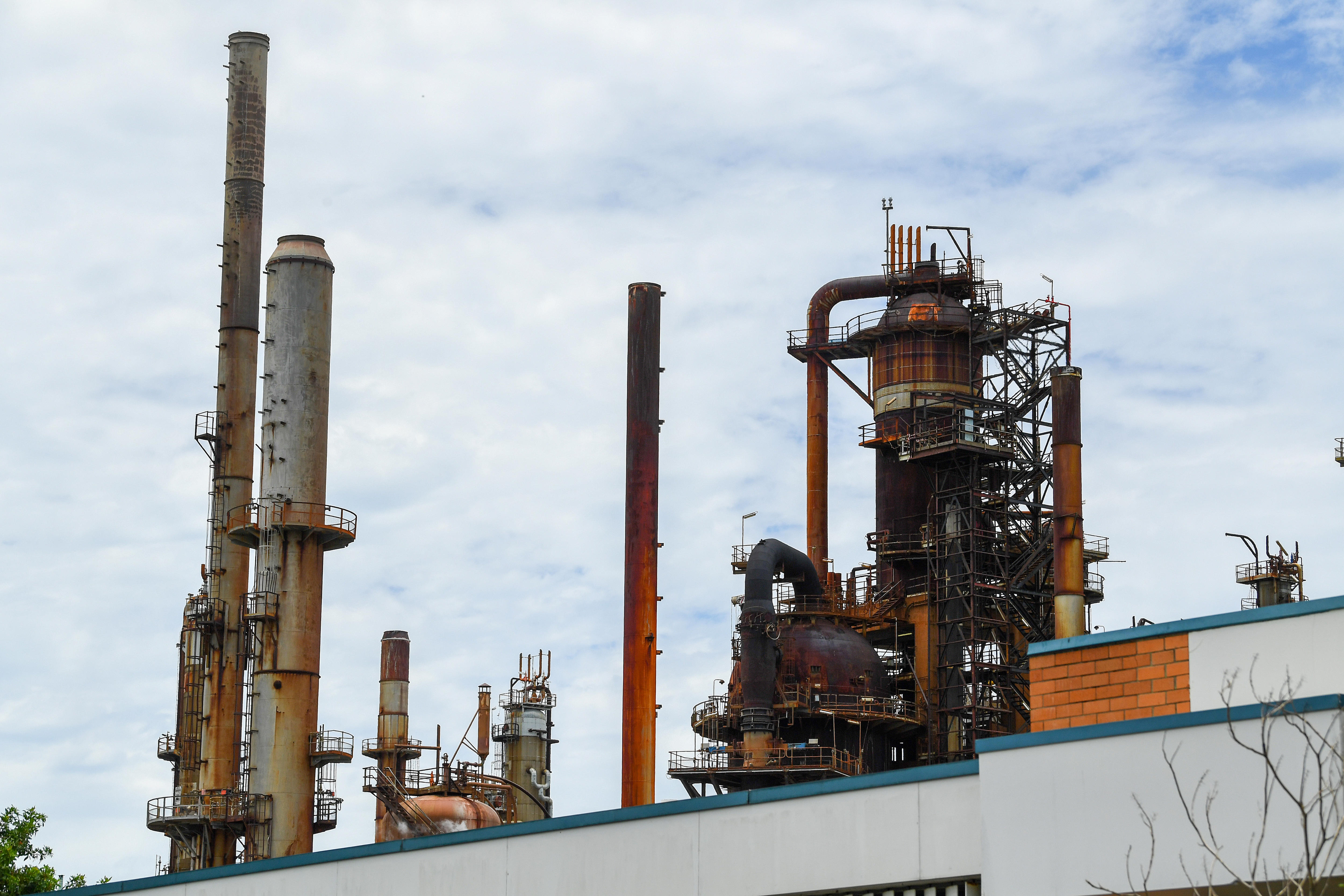 An older-looking oil refinery, with spires and chimneys, against a backdrop of a clouded sky.