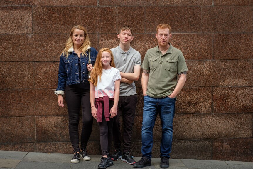 A woman, young girl, teenage boy, and man pose against an urban wall